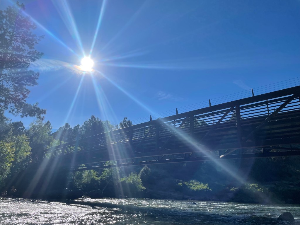 Image of sun and beams of light streaming through blue sky above a wooden bridge at First Street Rapids along the Deschutes River. Sunshine is reflecting off water below bridge and tree are along the riverbank.